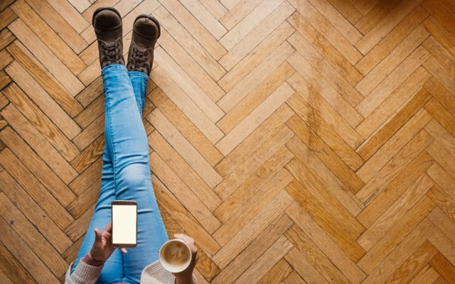 Mensch sitzt auf Fischgrät-Holzboden mit Handy und Kaffee