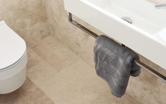 Helle Natursteinfliesen neben einer Badewanne mit grauem Handtuch